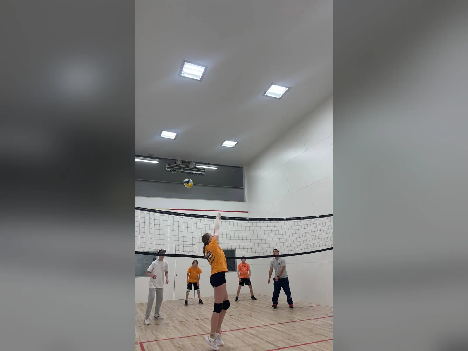 Підлітка подає волейбольний м’яч, поки партнери по команді й тренери спостерігають у спортивній залі.