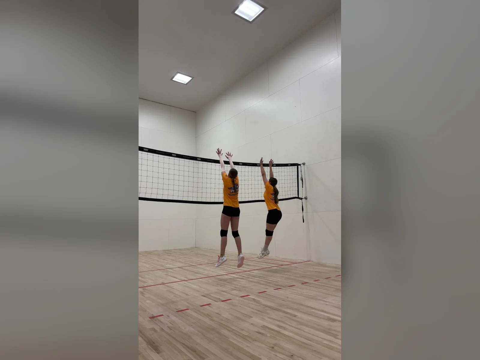 Дві юні волейболістки разом стрибають біля сітки під час тренування.